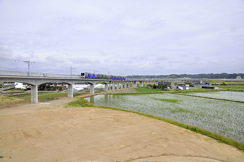 タイトル写真の仙石線 野蒜-陸前小野間のお立ち台から、反対向き石巻方の新線切り替え地点の眺め。HB-E210系気動車がいる少し右寄りから新線であるが、途中から高架の橋脚のタイプが変わっているのは、おそらく右は旧高架線の基礎を流用しているためであろう。そしてさらにその右奥の堤防の先に架かるのは鳴瀬川橋梁で、2000年6月25日に架け替えられており、それ以前には少し上流(写真では左)に架かっていた。ちなみにこの鳴瀬川橋梁はプレストレスト・コンクリート構造下路桁のフィンバック形式という構造をしており、1999年度に土木学会田中賞・日本コンクリート工学協会作品賞、2001年度に土木学会デザイン賞優秀賞を受賞している。