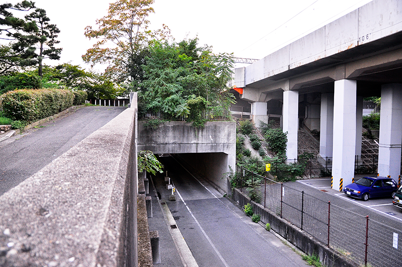 石屋川東岸の土手を住吉駅側から眺めたところ。高架線柱の左1本目と2本目の間に上写真の石碑が見える。なお、道路は現在も川を水底トンネルでくぐっている。