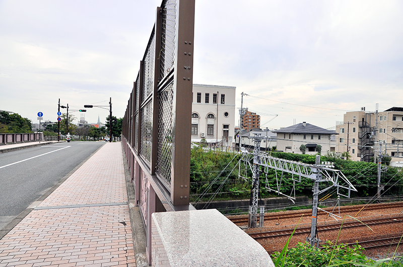 芦屋川に並行する道路を北西側から眺めたところ。中央のビルが上写真などに出てくるベージュの建物で、左隅の欄干のさらに左に芦屋川が流れている。