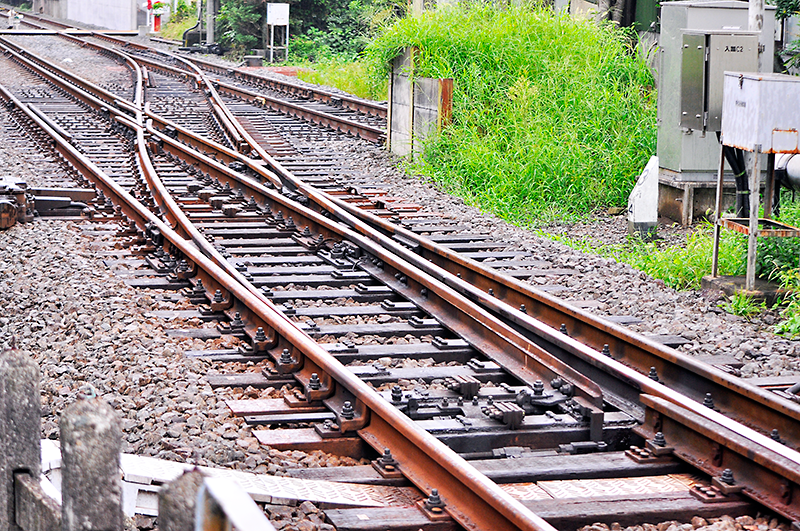 上写真の3線軌条の分岐器を箱根湯本駅寄りから入生田駅構内方を眺めたところ。この写真では曲線側が開通している状態。1,067mmゲージのみレールが、1,435mmゲージのみレールと交差する部分には可動クロッシングが使用されている。曲線側1,067mmゲージから3線軌条への合流では、車輪が固定クロッシング2つ、可動クロッシング、トングレールの順に通ってゆくため、3線軌条の分岐器らしい音が聴ける。