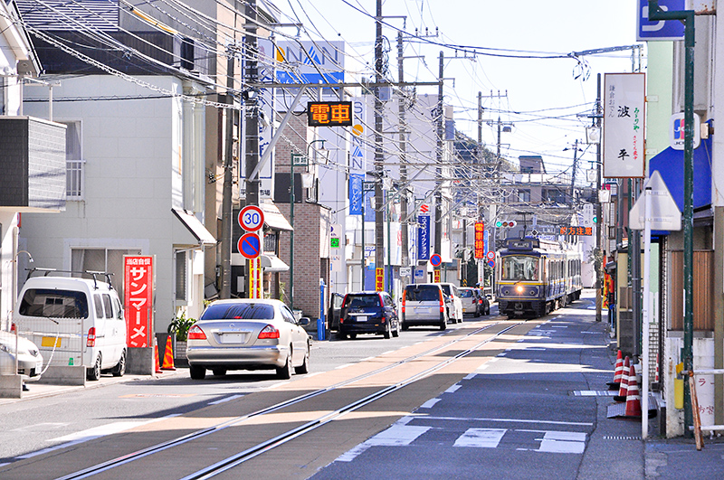 腰越-江ノ島 間の併用軌道。商店街の歩道がない道路の真ん中に単線の併用軌道が敷かれている、それだけで面白い光景だが、そこを通るのが鉄道車輛という対比が、これまた面白い。写真は江ノ島駅寄りから腰越駅方の眺め。車輛は10形電車。