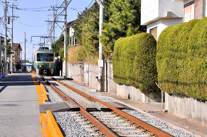 七里ヶ浜-鎌倉高校前 間の併用軌道で、七里ヶ浜駅寄りから鎌倉高校前駅方を眺めたところ。縁石は黄色く塗られているとはいえ、柵とかはない。しかしそれが「玄関先を通る」江ノ電らしい光景なのだともいえる。車輛は500形電車。