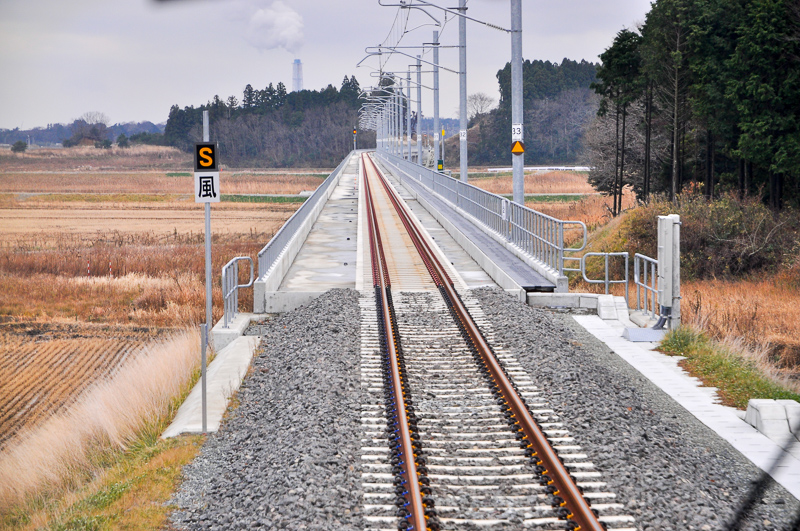 坂元駅から駒ヶ嶺方に約5km程の地点にある切り通しから高架線に切り替わる地点。この写真を掲載した理由は、次からでこの辺の建設中の写真を紹介しているからになる。なお、奥に見える煙突は相馬火力発電所のモノ。