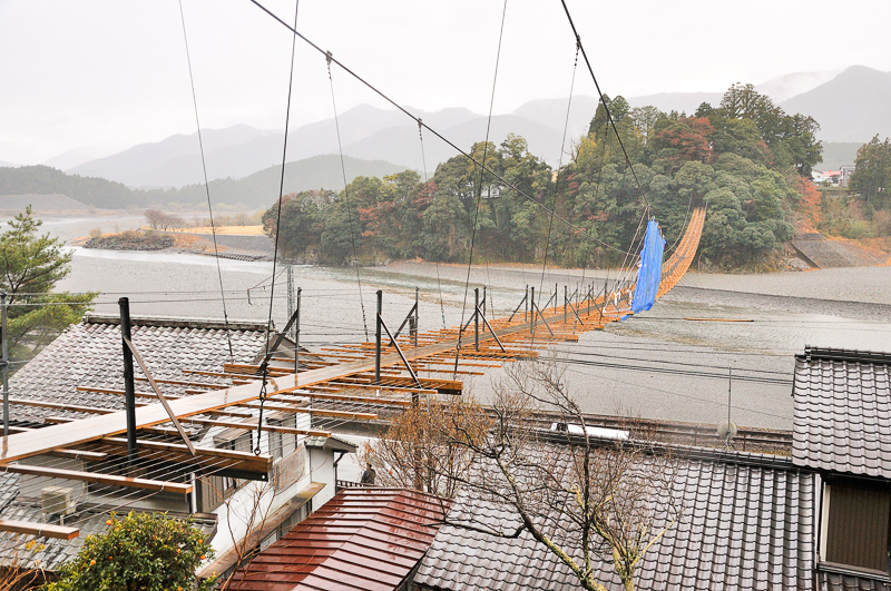 東岸袂付近千頭側から眺めた吊り橋。民家の屋根の上空や県道、線路の上をも通っているのが見てとれる。